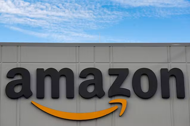 The official corporate logo of Amazon displayed on a fulfillment building exterior sign against a blue sky.